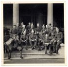 Photograph of officers of the 1st Battalion, in party hats, and smoking cigars, seated on steps outside the front entrance of the officers' mess, Shanghai, China, n.d., [winter 1937 - 1939]