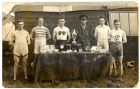 Photograph of the prizewinners in a foot runners competition, standing behind a table of trophies, including, second from right, Bugler Sammy Lee of No. 3 Company, 5th Battalion The Durham Light Infan