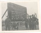 Photograph of soldiers [possibly of the 113 Light Anti-Aircraft Regiment, Royal Artillery (Territorial Army)] standing beside a temporary memorial plaque at Belsen Concentration Camp, taken in Germany