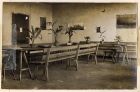 Photograph of a room with benches and trestle tables with assorted plants, captioned: VII. The 'silence' room, about the noisiest in camp, German papers, games, communiques etc., were obtainable here,